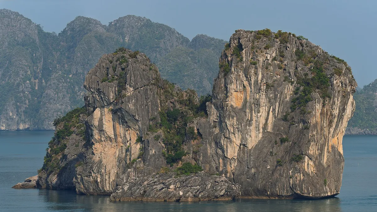 A sheer limestone island rising from the still water of Ha Long Bay, with hazy karst hills behind