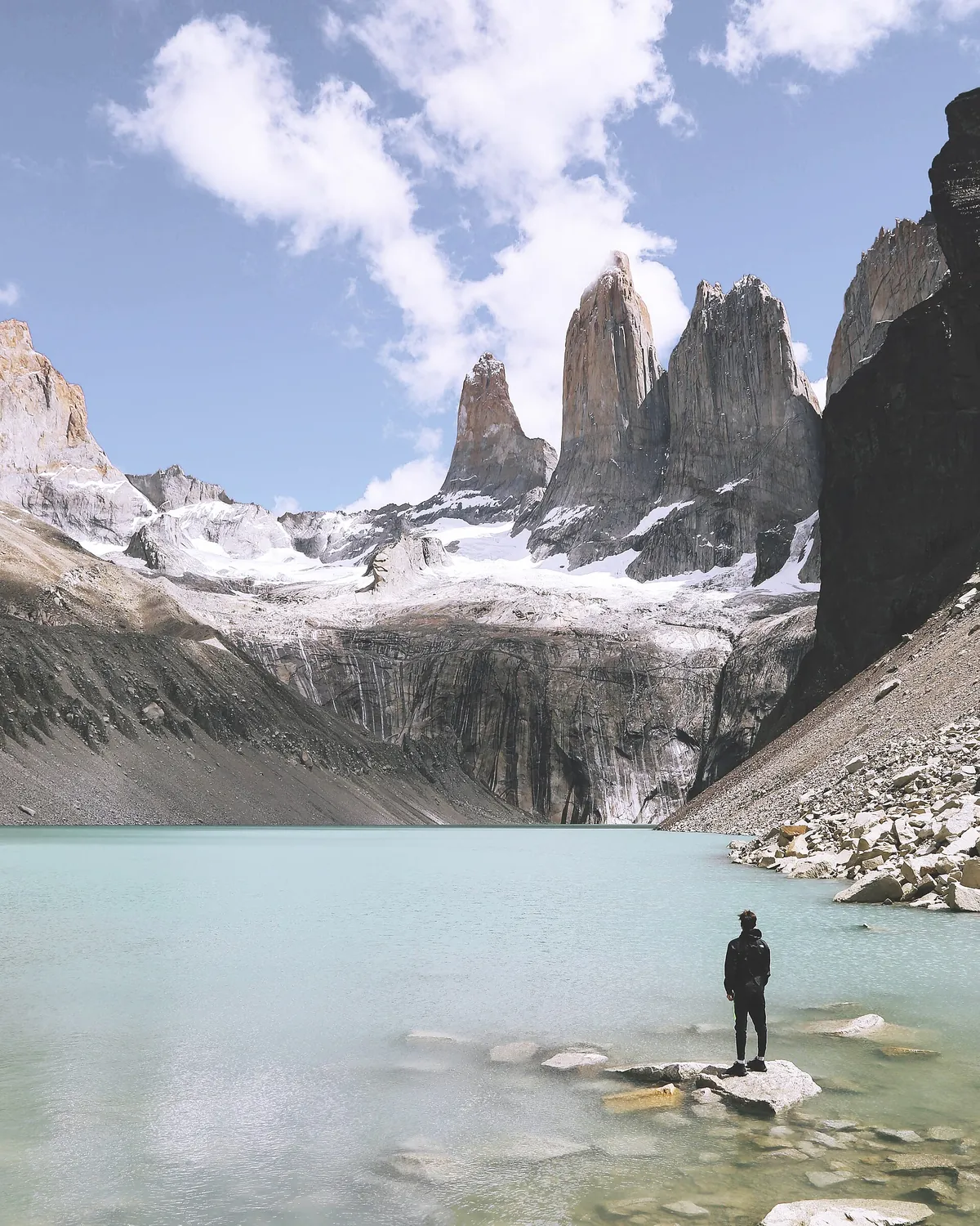 A hiker standing on a rock in a pale green glacial lake below the granite towers of Torres del Paine